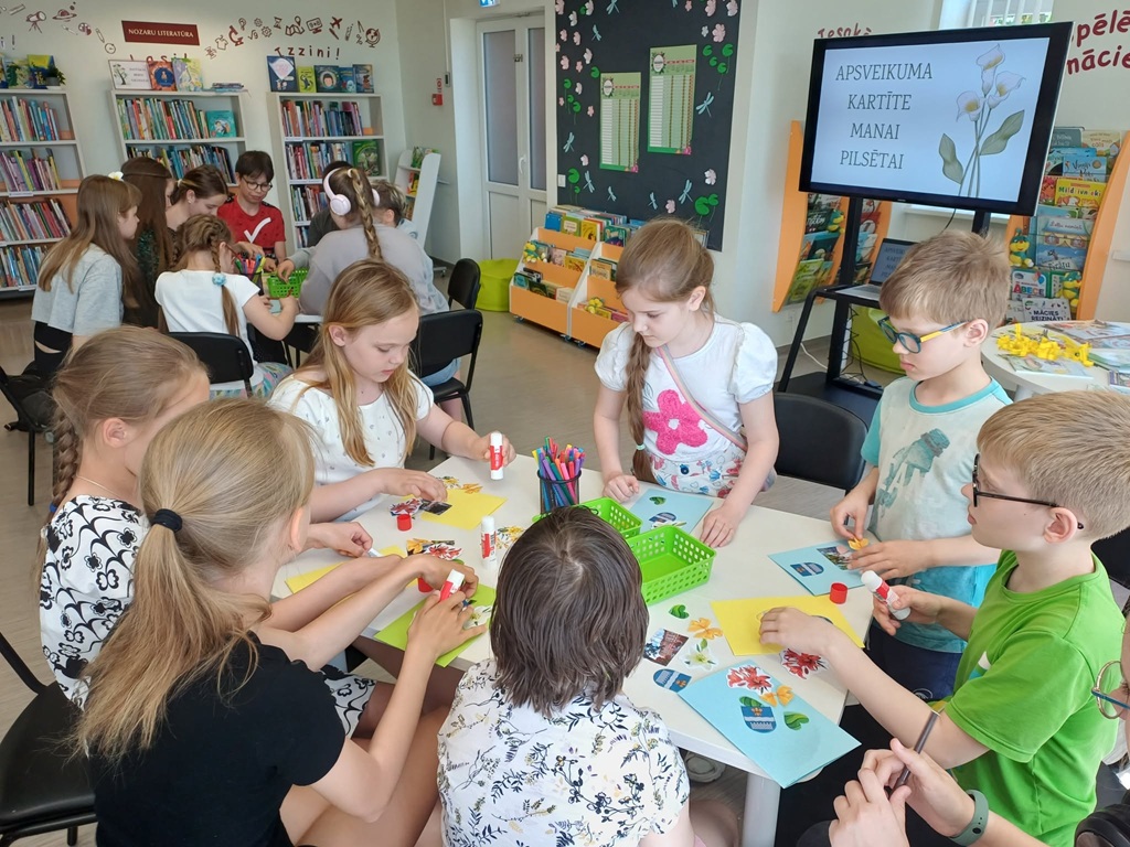 Bērnu grupa bibliotēkas telpā sēž ap apaļu galdu un radošā nodarbībā veido apsveikuma kartītes, izmantojot līmi, flomasterus un krāsainus papīrus. Fonā pie sienas redzams TV ekrāns ar uzrakstu 'APSVEIKUMA KARTĪTE MANAI PILSĒTAI' un grāmatu plaukti."