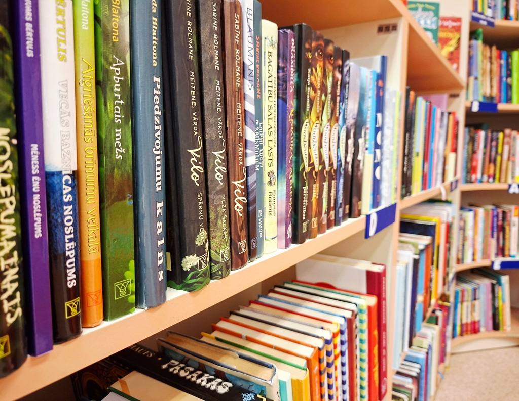 Grāmatu plaukti, kas piepildīti ar krāsainām, vertikāli sakrautām grāmatām bibliotēkā. Uz muguriņām attēloti dažādi nosaukumi dažādās valodās, rosinot ziņkāri un rosinot izpēti.