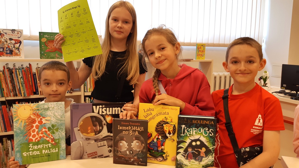 Četri bērni stāv bibliotēkā pie galda un rāda dažādas grāmatas, starp kurām ir 'Žirafīte steidz palīgā', 'Visums', 'Izmeklētāji no 3. A', 'Labu labā lasāmgrāmata' un 'Ikabogs'. Meitene fonā uz augšu pacēlusi zaļu papīra lapu ar uzrakstu 'Grāmatu Bingo'.