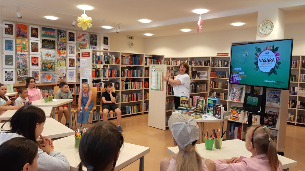 asākuma vadītāja stāv bibliotēkas telpas vidū un uzrunā bērnus, kuri sēž uz krēsliem pie galdiem. Labajā pusē redzams liels ekrāns ar pasākuma afišu 'SKAISTĀ VASARA BIBLIOTĒKĀ', bet fonā izvietoti grāmatu plaukti un pie sienas piestiprināti bērnu zīmējumi.