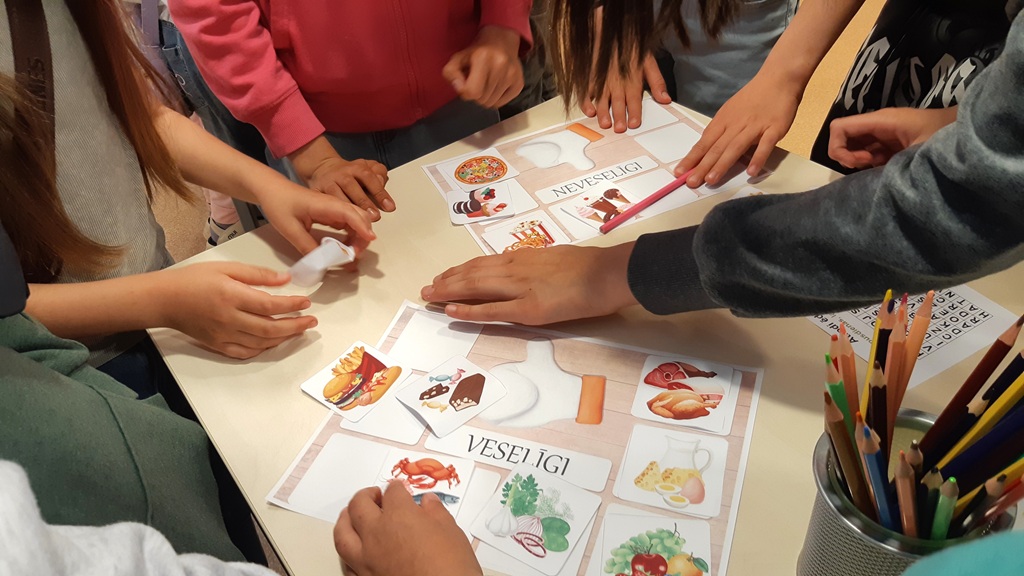 Tuvplāns no augšas: bērnu rokas saliek izglītojošu spēli uz galda, šķirojot pārtikas produktu attēlus. Uz vienas pamatnes ar uzrakstu 'VESELĪGI' izvietotas kartītes ar dārzeņiem, zivīm un piena produktiem, bet uz otras ar uzrakstu 'NEVESELĪGI' – ar picu, saldumiem un frī kartupeļiem. Priekšplānā redzams zīmuļu trauks.