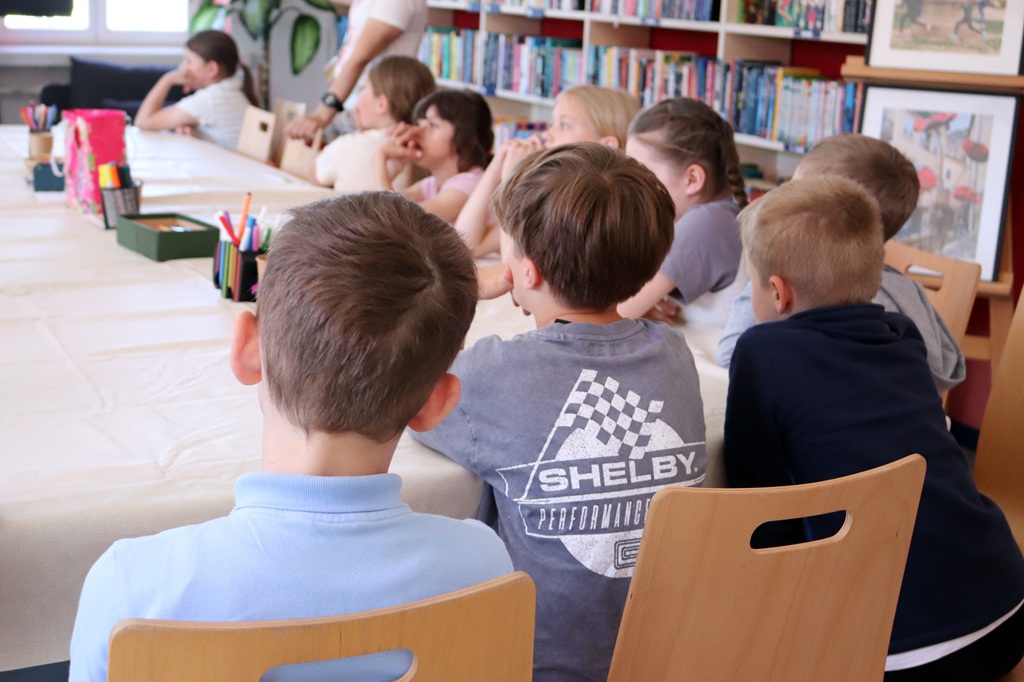 Bērni sēž ap galdu bibliotēkā un uzmanīgi kaut ko vēro. Redzami grāmatu plaukti un mākslas darbi rada siltu, izglītojošu atmosfēru.