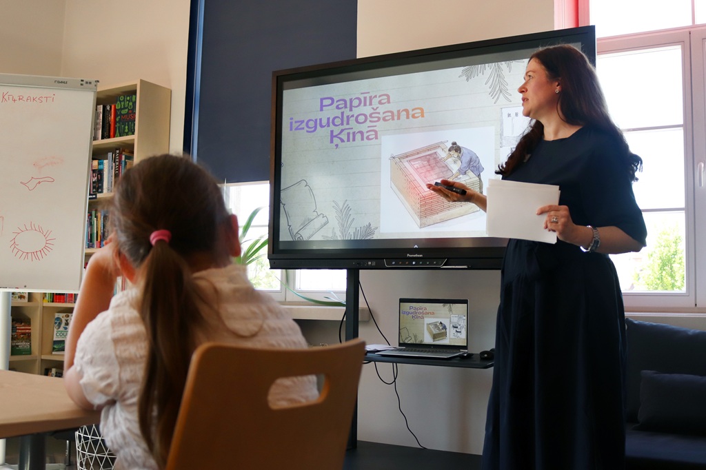 Sieviete klasē rāda uz ekrānu, kurā redzams teksts par papīra izgudrošanu. Jauna meitene uzmanīgi vēro. Aina šķiet izglītojoša.
