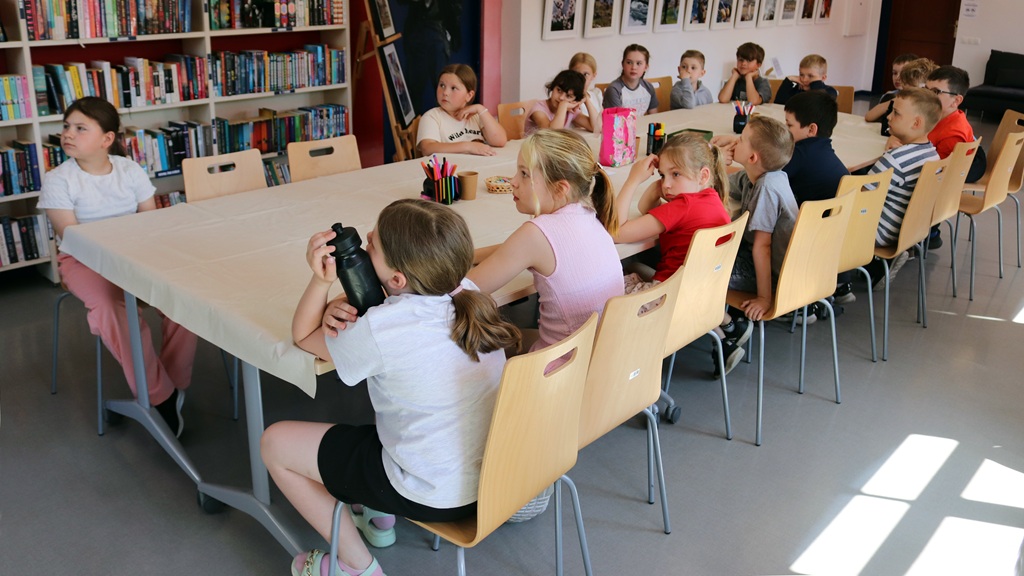 Bērnu grupa uzmanīgi sēž ap lielu galdu bibliotēkā, ko ieskauj grāmatu plaukti. Viņi šķiet iesaistīti un zinātkāri, uzmanīgi klausoties.