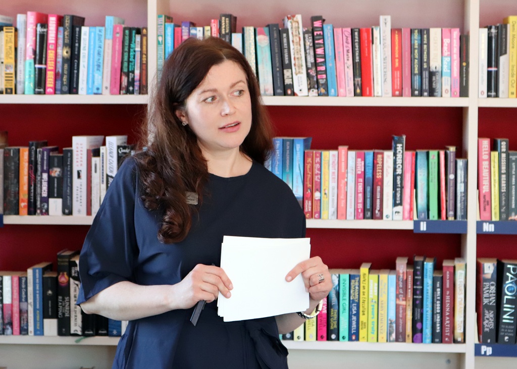 Sieviete ar gariem brūniem matiem tur rokās papīrus un runā krāsaina grāmatu plaukta priekšā. Viņa izskatās domīga un aizņemta bibliotēkas vidē.