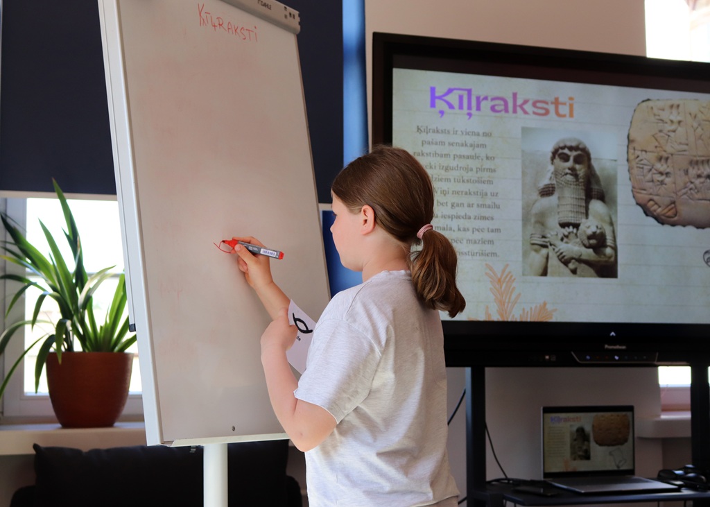 Jauna meitene raksta uz tāfeles klasē. Fonā esošais digitālais ekrāns rāda senus tekstus un attēlus. Telpā valda gaiša, akadēmiska atmosfēra.