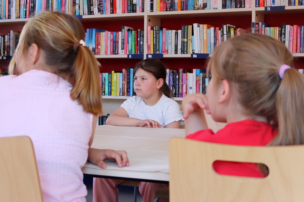 Trīs bērni sēž pie galda bibliotēkā. Meitene baltā tērpā skatās uz priekšu, izskatoties domīga. Fonā ir grāmatu plaukti, kas piepildīti ar krāsainām grāmatām.