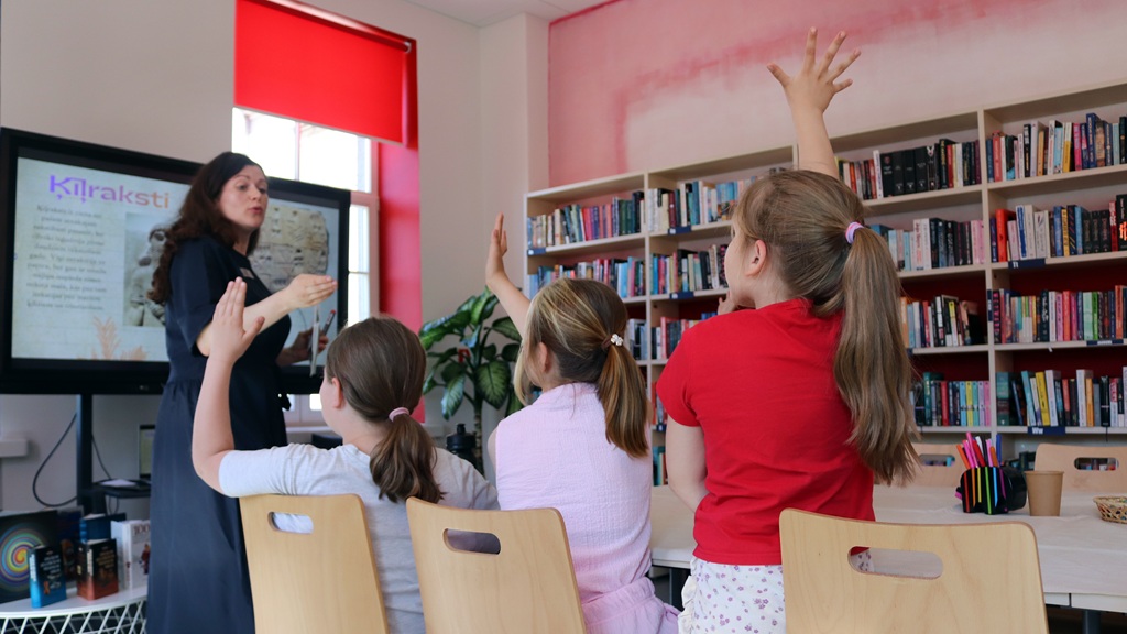 Skolotājs klasē stāv pie displeja ekrāna un sarunājas ar trim skolēniem, kas paceļ rokas. Grāmatu plaukti atgādina bibliotēkas vidi.