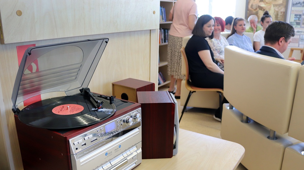 Tuvplāns bibliotēkā: priekšplānā uz galda novietots retro stila plašu atskaņotājs ar koka apdari, kurā griežas vinila plate. Fonā uz dīvāniem un krēsliem sēž pasākuma dalībnieki.