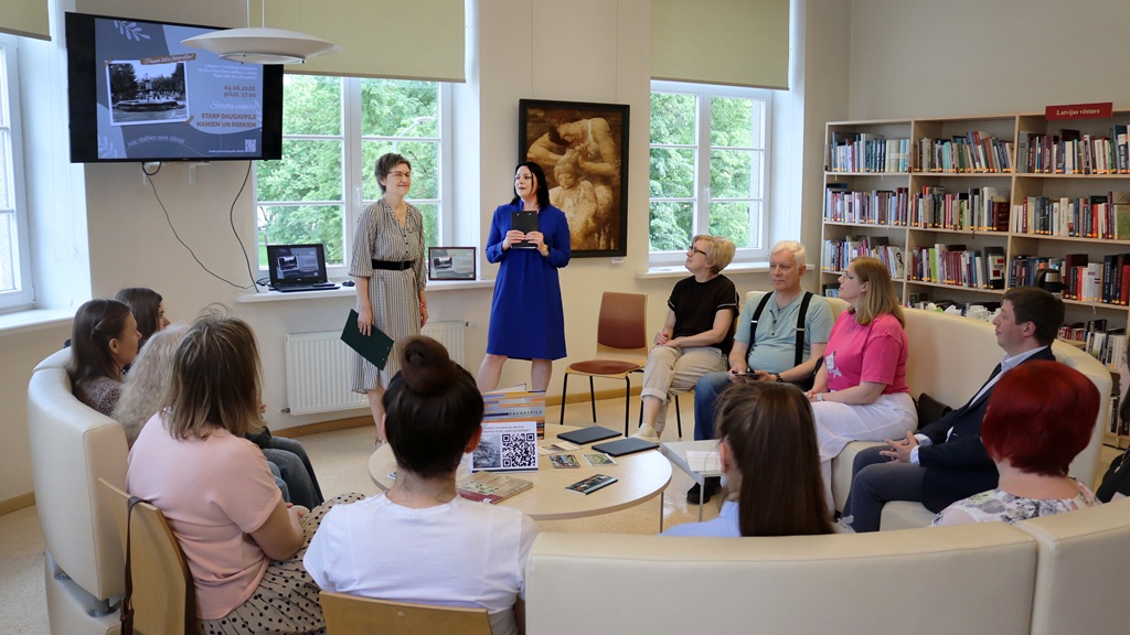 Kopskats no aizmugures uz pasākumu bibliotēkā: divas vadītājas stāv telpas priekšā pie liela ekrāna, uz kura redzams afišas slaids. Klausītāji sēž puslokā uz gaišiem dīvāniem un krēsliem.