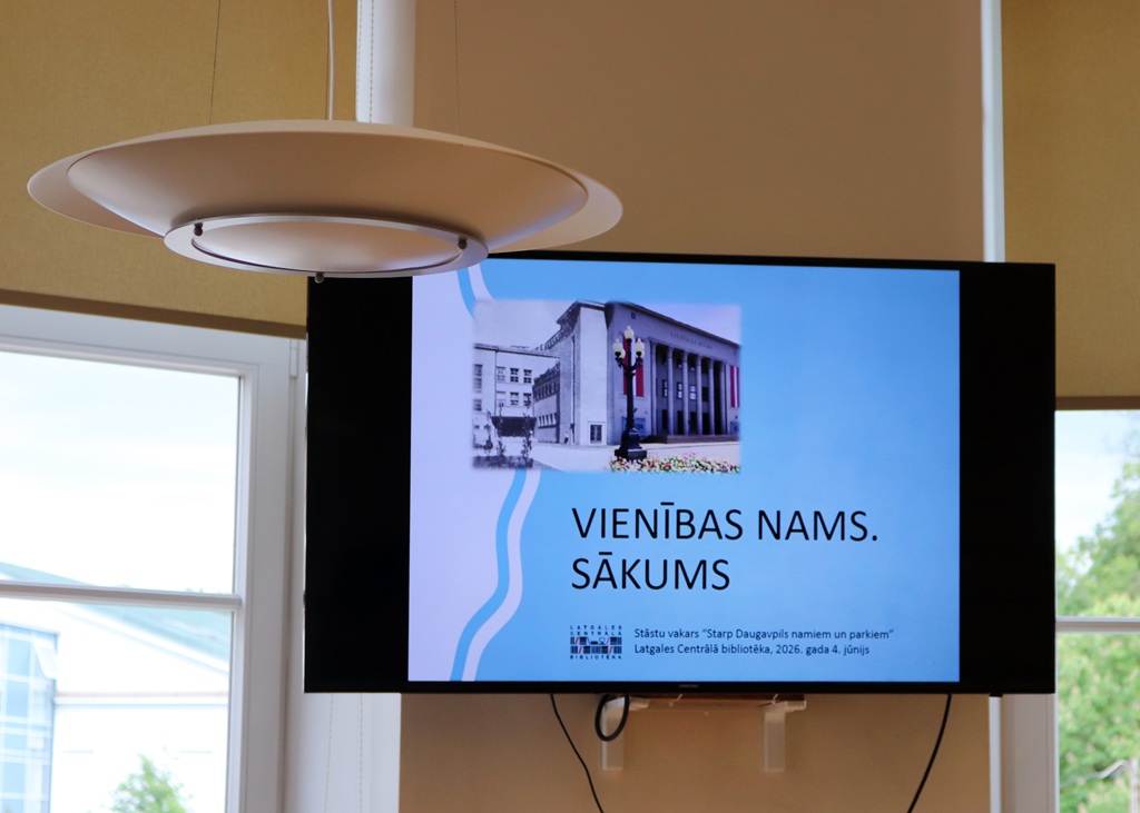 Pie sienas piestiprināts liels plakanā ekrāna televizors, kurā redzams prezentācijas slaids ar Vienības nama attēlu un uzrakstu "VIENĪBAS NAMS. SĀKUMS". Virs televizora redzama moderna dizaina griestu lampa.