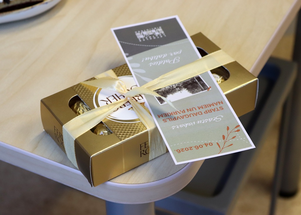 Tuvplāns no augšas: uz gaiša koka galda stūra novietota zeltīta 'Ferrero Rocher' konfekšu kārba ar krēmkrāsas lenti. Tam virsū ir piesprausta zaļgana dizaina kartīte ar uzrakstu 'Paldies par dalību!