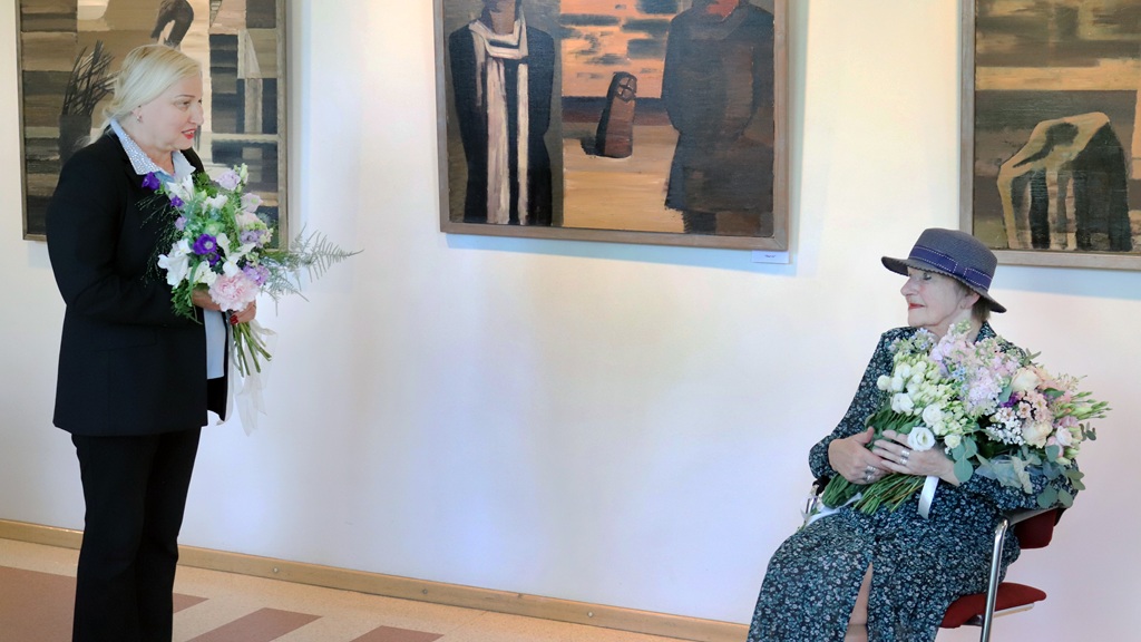 Divas sievietes galerijā, viena stāv ar ziediem, otra sēž, smaida un tur ziedus. Gleznojumi pie sienas liek domāt par mākslas izstādi.
