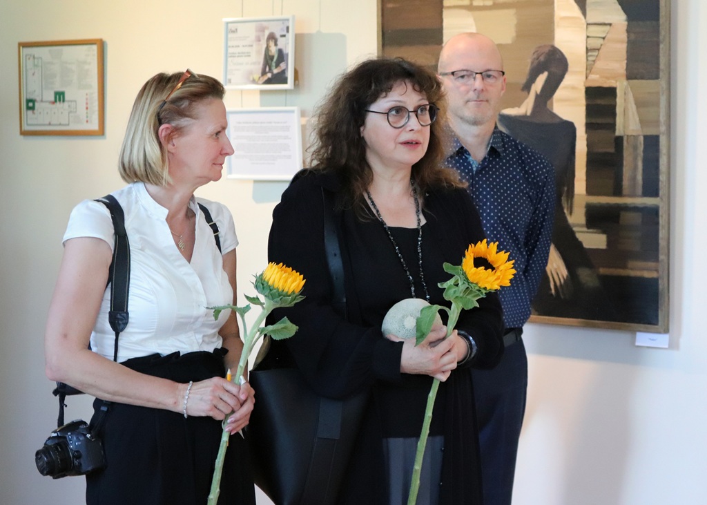 Trīs cilvēki stāv mākslas galerijā. Divas sievietes tur saulespuķes, un vīrietis stāv aiz viņām. Fonā redzama glezna. Atmosfēra šķiet pārdomīga un svinīga.