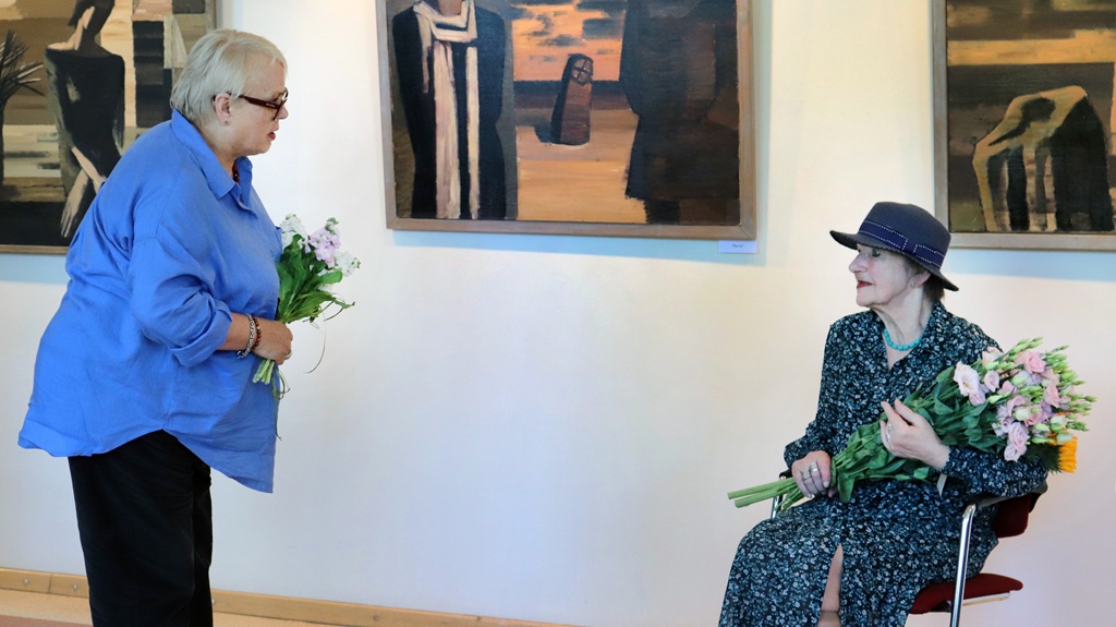 Divas vecāka gadagājuma sievietes mākslas galerijā apmainās ar ziediem. Viena stāv zilā blūzē un pasniedz ziedus, bet otra sēž cepurē un rakstainā kleitā, smaidot. Pie sienas redzamas gleznas.