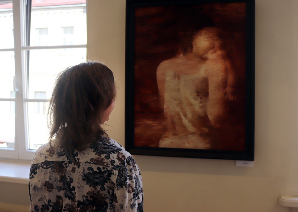 Cilvēks ar pleciem gariem matiem rakstainā kreklā stāv pie gleznas galerijā. Mākslas darbā attēlota izplūdusi figūra ar bērnu. Aina ir mierīga un pārdomu pilna.