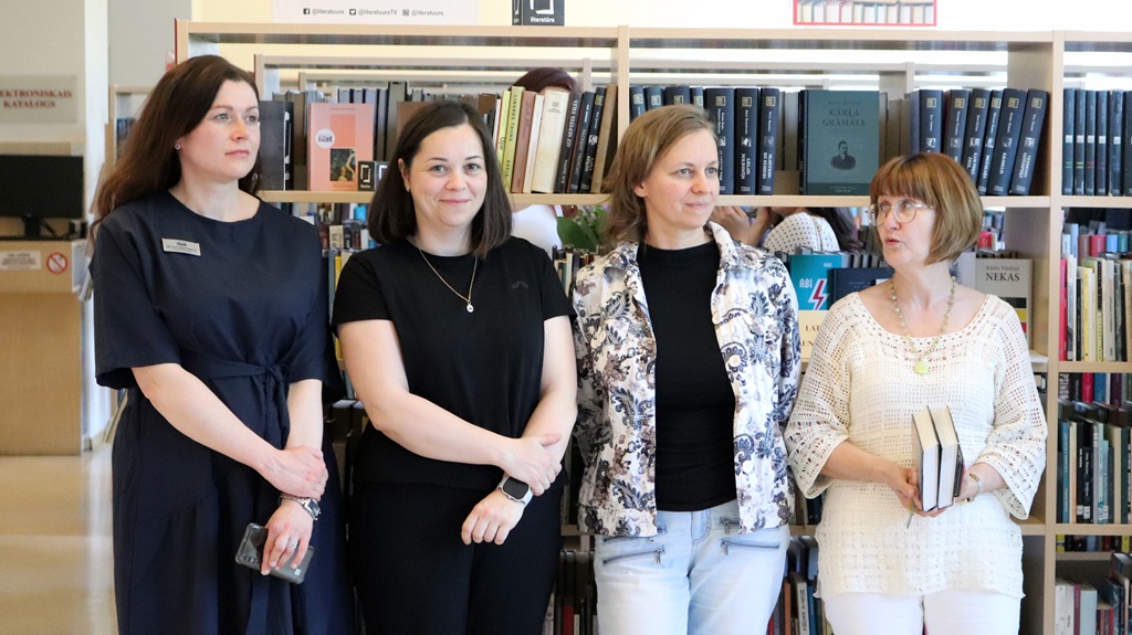 Četras sievietes stāv bibliotēkas grāmatu plaukta priekšā, šķietami iesaistījušās un koncentrējušās. Viena tur rokās grāmatas, kas norāda uz literāru vai akadēmisku notikumu.