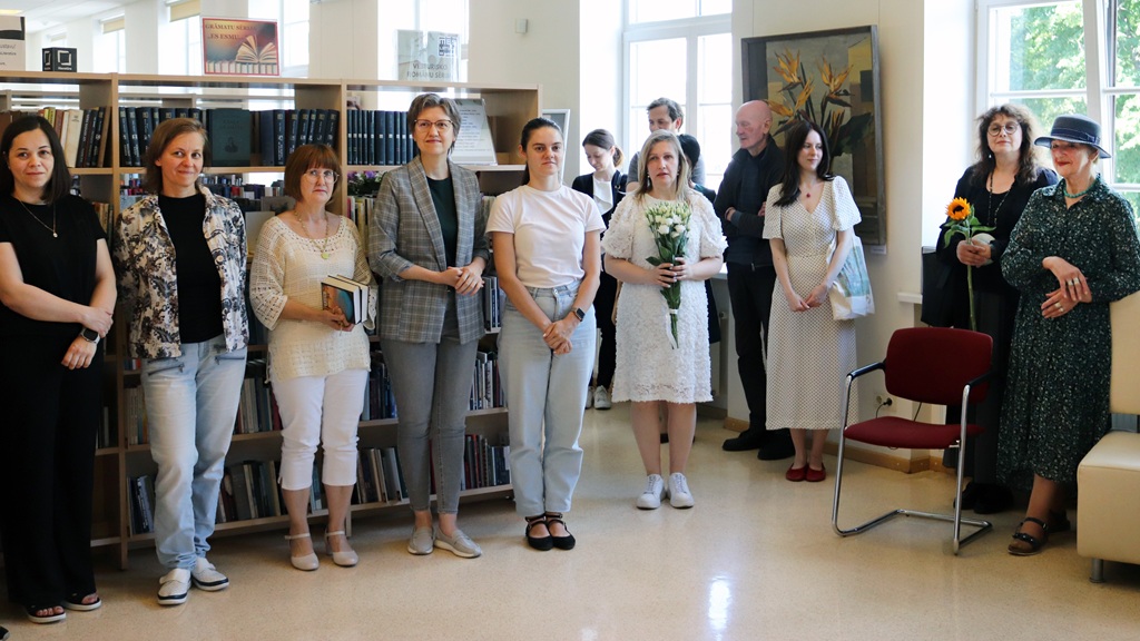 Cilvēku grupa uzmanīgi stāv bibliotēkā, fonā grāmatu plaukti. Daži tur ziedus, radot siltu un svinīgu atmosfēru.