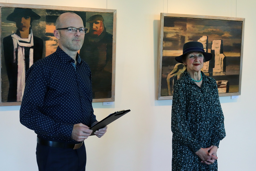 Vīrietis brillēs, turot rokās planšeti, stāv blakus smaidošai sievietei cepurē. Viņi atrodas mākslas galerijā, fonā divas abstraktas gleznas.