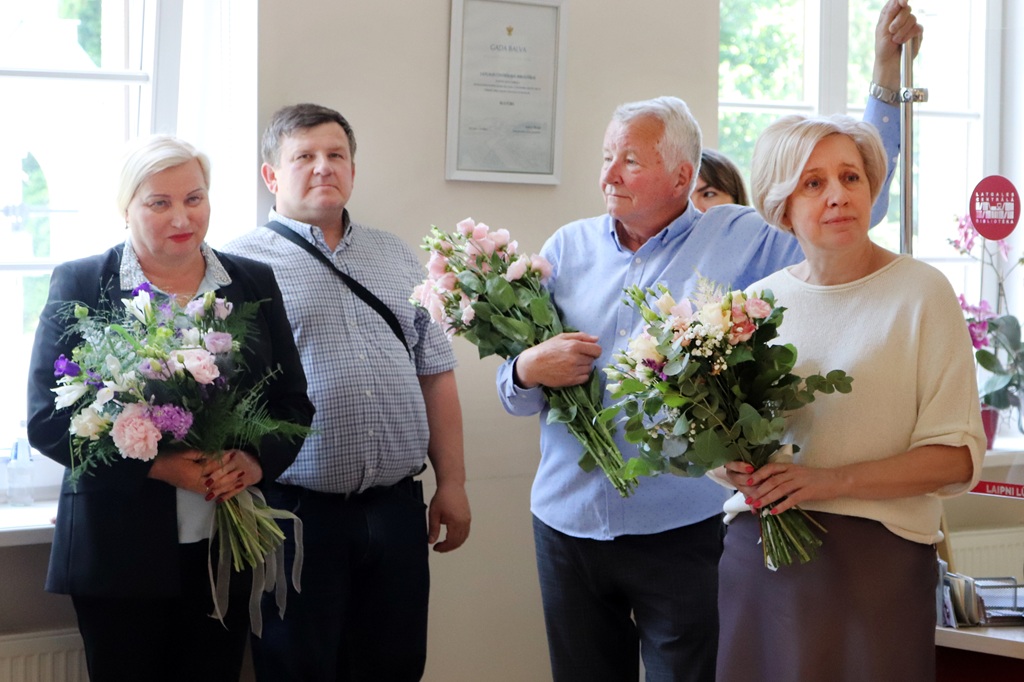 Četri cilvēki stāv iekštelpās, katrs turot ziedu pušķi. Šķiet, ka viņi svin vai apmeklē īpašu pasākumu. Noskaņojums ir silts un svētku pilns.