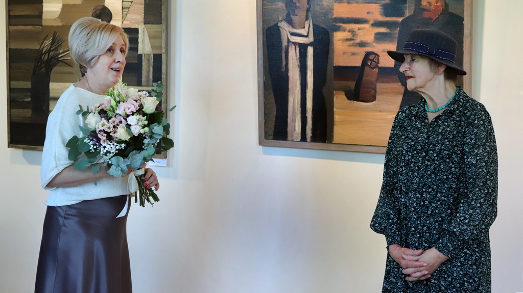 Divas sievietes stāv mākslas galerijā. Viena tur ziedu pušķi un smaida, bet otra, cepurē galvā, klausās. Aiz viņām pie sienas karājas gleznas.