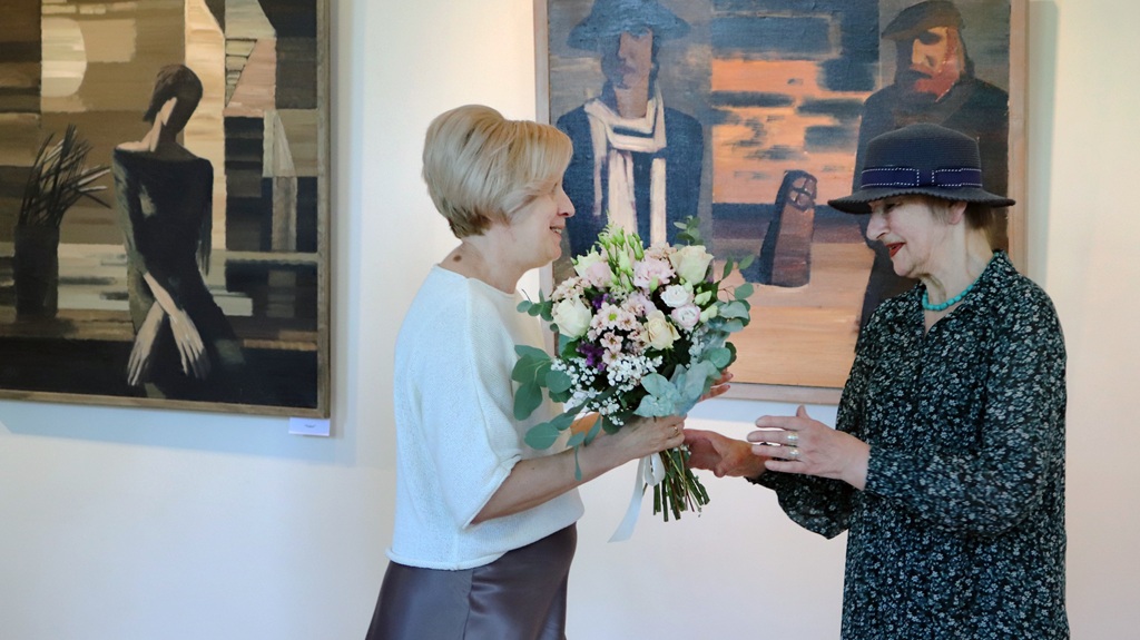 Divas sievietes apmainās ar pušķi mākslas galerijā. Vienai mugurā ir rakstaina kleita un cepure, otrai – balta blūze. Fonā gleznas pieklusinātās krāsās.