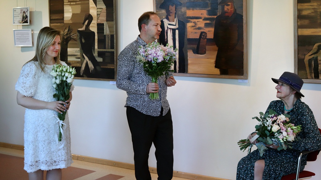 Jauna sieviete un vīrietis, abi turot rokās ziedu pušķus, stāv blakus vecāka gadagājuma sēdošai sievietei ar ziediem. Aiz viņiem pie galerijas sienas karājas mākslas gleznas.