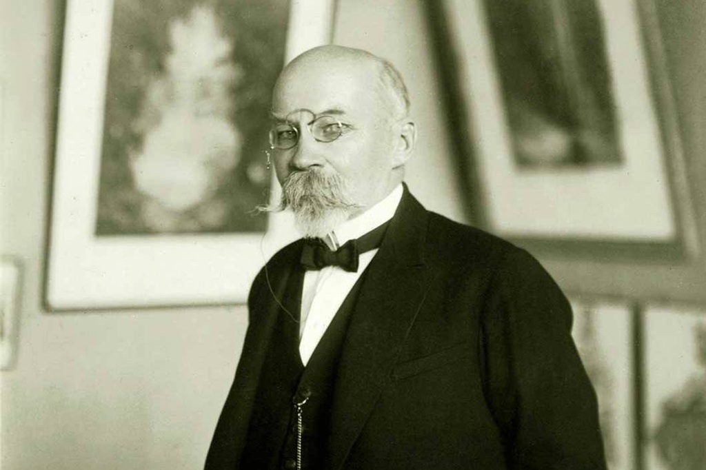 Gados vecāks vīrietis ar monokli un ūsām, uzvalkā un tauriņā, stāv izplūdušu ierāmētu mākslas darbu priekšā. Tonis ir formāls un akadēmisks.