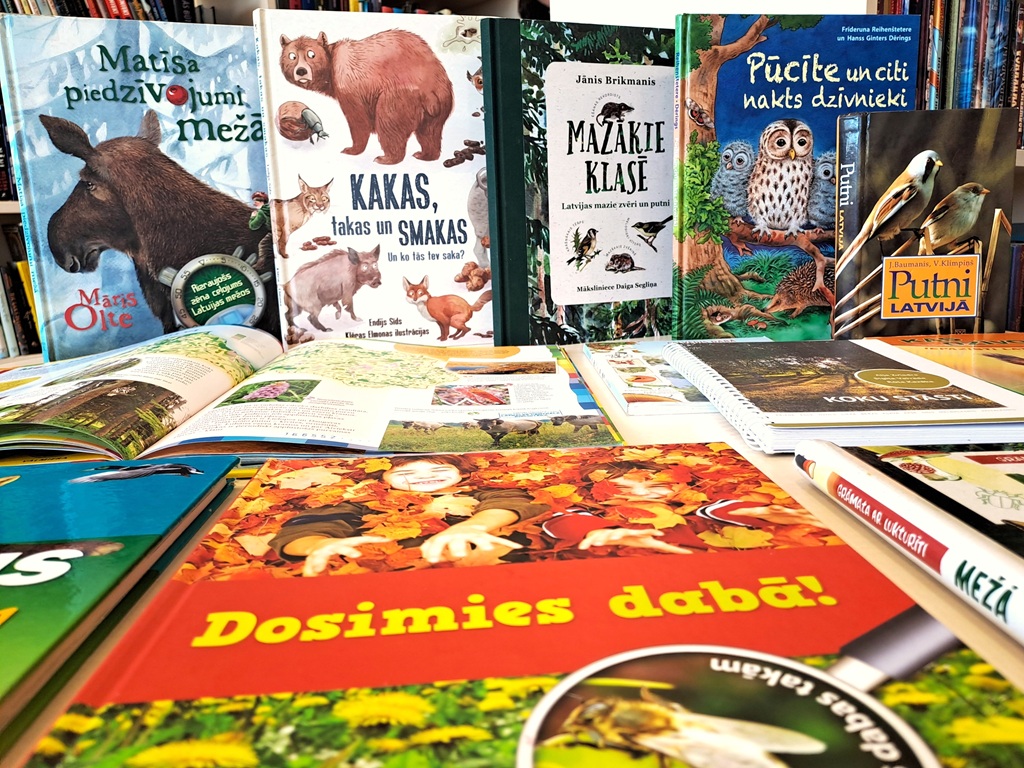 Dažādas ilustrētas grāmatas par mežu, dabas pētīšanu un dzīvniekiem, piemēram, "Matīsa piedzīvojumi mežā" un "Pūcīte un citi nakts dzīvnieki", izkārtotas stateniski un guļus uz galda, simbolizējot aizraujošu ceļojumu meža pasaulē.