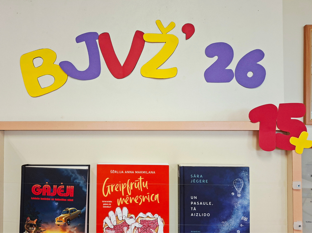Grāmatu izstādes stends ar uzrakstu "BJVŽ 26" un stateniski trīs plaukta izvietotām grāmatām: "Gājēji", "Greipfrūtu mēnesnīca" un "Un pasaule, tā aizlido".