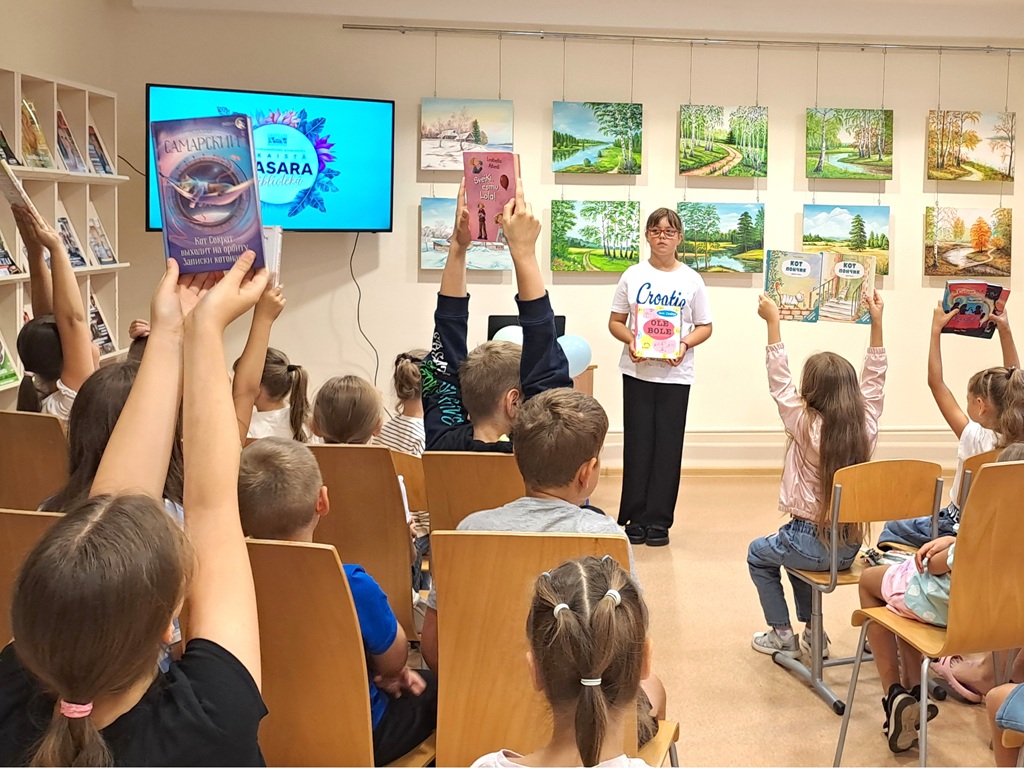 Pasākuma dalībnieki bibliotēkas zālē ar paceltām grāmatām, fonā redzami mākslas darbi