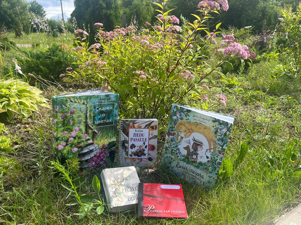 Piecas dažādas grāmatas par ziediem un dabu novietotas uz zāles dārza fonā, kurā aug ziedošs krūms ar rozā ziedkopām.