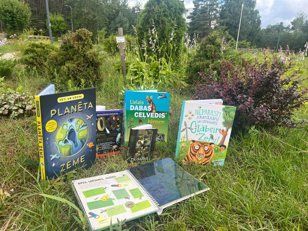 Dabas izziņas grāmatas izkārtotas zaļā zālienā dārza fonā. Starp grāmatām ir arī atvērts izdevums par kukaiņiem.