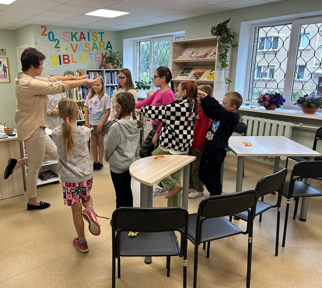 Bibliotekāre un bērnu grupa sastājušies aplī, kopā piedalās kustību rotaļā, nostājoties uz vienas kājas un izstiepjot rokas.