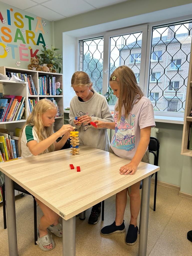 Trīs meitenes bibliotēkā aizrautīgi spēlē galda spēli, kārtojot koka torņa klucīšus uz galda.