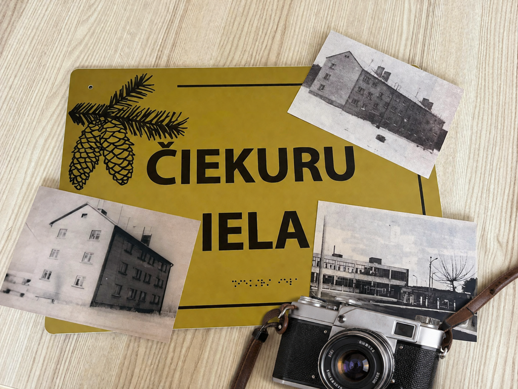 Tematisks attēls uz koka virsmas, kas reklamē fotoorientēšanās spēli "Čiekuru ielā". Tajā redzama dzeltena ielas plāksne ar uzrakstu "ČIEKURU IELA" un čiekuru attēlu. Blakus novietota sena, sudrabaina fotoaparāta kamera ar ādas siksniņu. Ap plāksni un kameru izkārtotas divas melnbaltas fotogrāfijas, kurās redzamas Čiekuru ielas ēkas pirms daudziem gadiem.