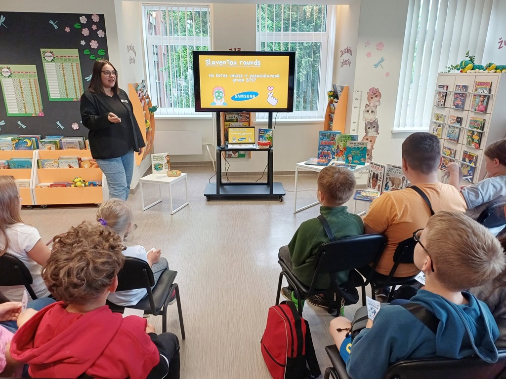 Bibliotekāre stāv pie TV ekrāna un vada viktorīnu bērnu auditorijai, kas sēž krēslos un klausās, vērojot prezentāciju ar uzrakstu "Slavenību raunds".