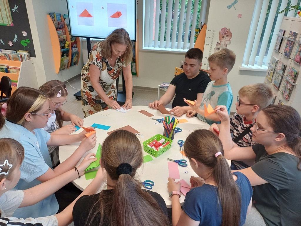 Meitenes un zēni sēž ap apaļu galdu bibliotēkā un kopā ar bibliotekāri locīšanas tehnikā veido papīra izstrādājumus jeb origami, turot rokās dažādu krāsu papīru.