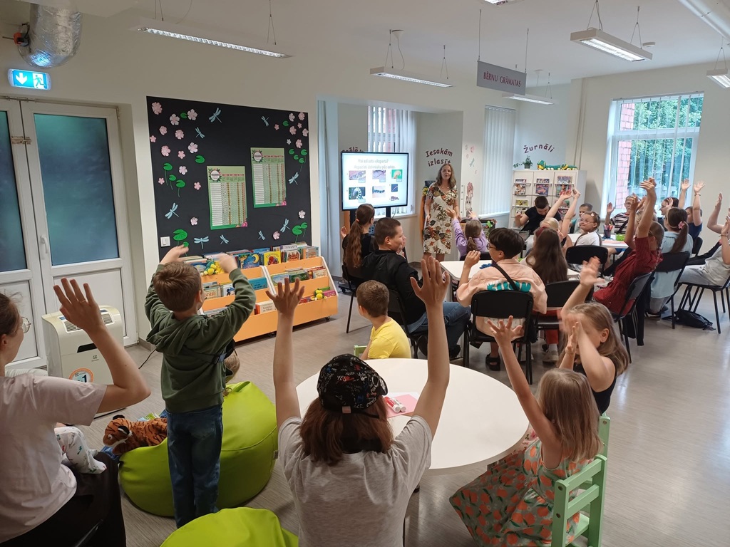 Bērnu grupa piedalās interaktīvā nodarbībā, sēžot pie galdiem un uz sēžammaisiem ar paceltām rokām, skatoties uz ekrānu un priekšā stāvošo bibliotekāri.