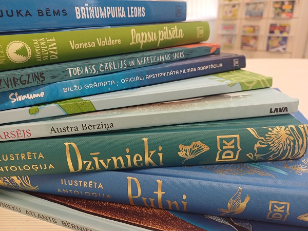 Grāmatu kaudze uz galda, kuras muguriņās redzami dažādi nosaukumi latviešu valodā, tostarp "Lapsu pilsēta", "Dzīvnieki", "Putni", "Sraume. Bilžu grāmata" un citi.