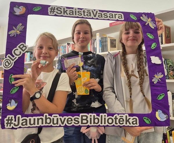 Trīs smaidīgas meitenes bibliotēkas telpā pozē, turot rokās lielu, purpursarkanu rāmi ar dekorācijām un uzrakstiem "Skaistā Vasara" un "Jaunbūves Bibliotēka".