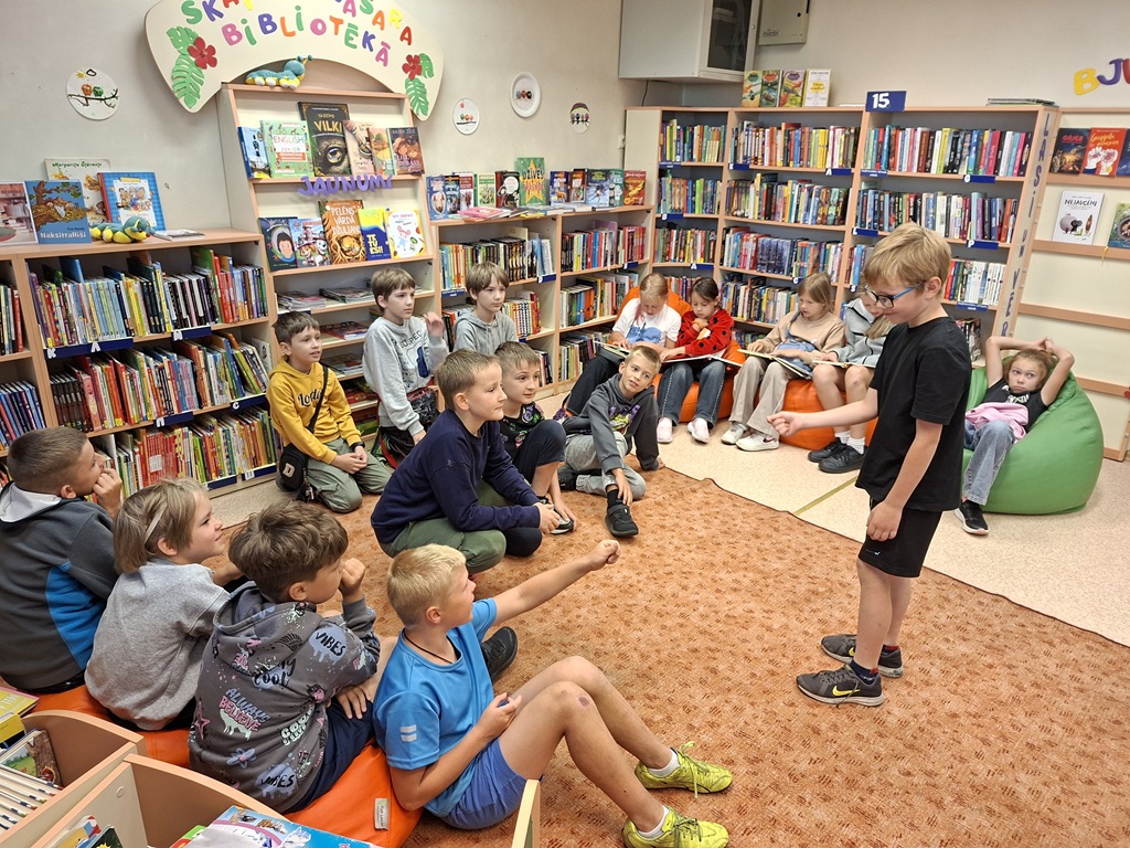 Bērnu grupa sēž bibliotēkā uz paklāja un sēžammaisiem, bet viens zēns stāv priekšplānā un ar rokām mēģina parādīt kādu vārdu, ko pārējiem bērniem ir jāatmin.