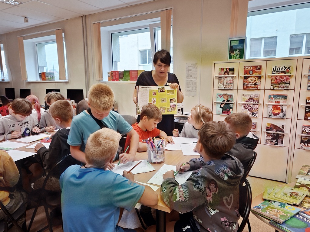 Bibliotekāre stāv pie galda un rāda bērniem atvērtu grāmatu, kamēr bērni sēž apkārt un zīmē vai klausās skatoties uz bibliotekāres rokās esošo grāmatu.
