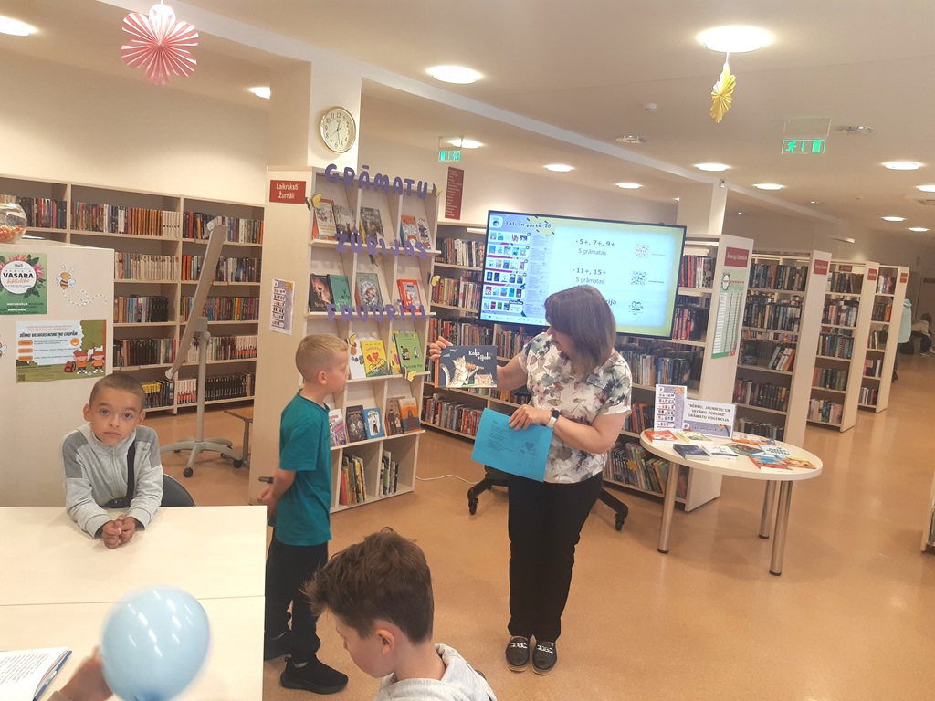 Bibliotekāre tur rokās grāmatu un rāda to bērniem, fonā redzami vairāki grāmatu plaukti un TV ekrāns ar prezentāciju.