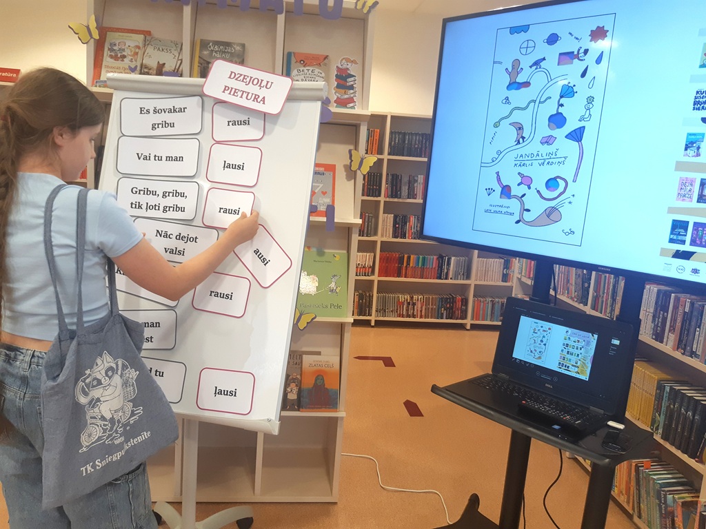 Meitene uz stenda "Dzejoļu pietura" kārto vārdus, piemēram, Es šovakar gribu, rausi, vai tu man, ļausi, , blakus stāv ekrāns un klēpjdators ar ilustrāciju "Jandāliņš Kārlis Vērdiņš"