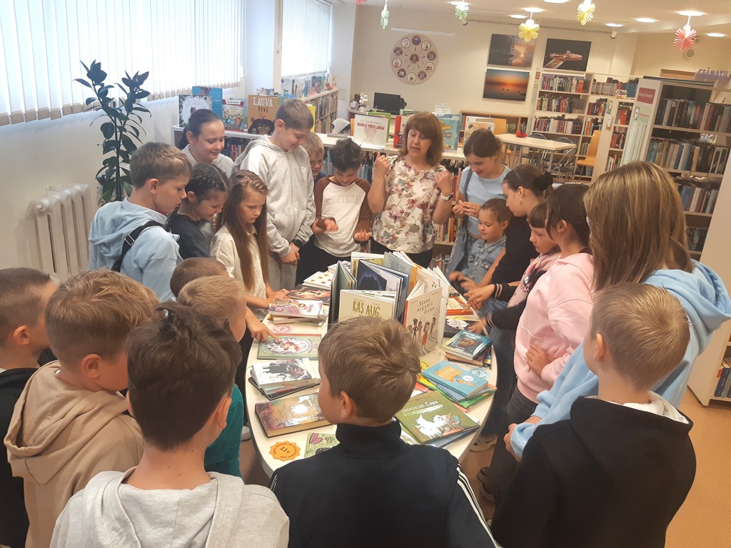 Bērnu grupa un bibliotekāre drūzmējas ap ovālu galdu, pētot un aplūkojot tur izliktās grāmatas.