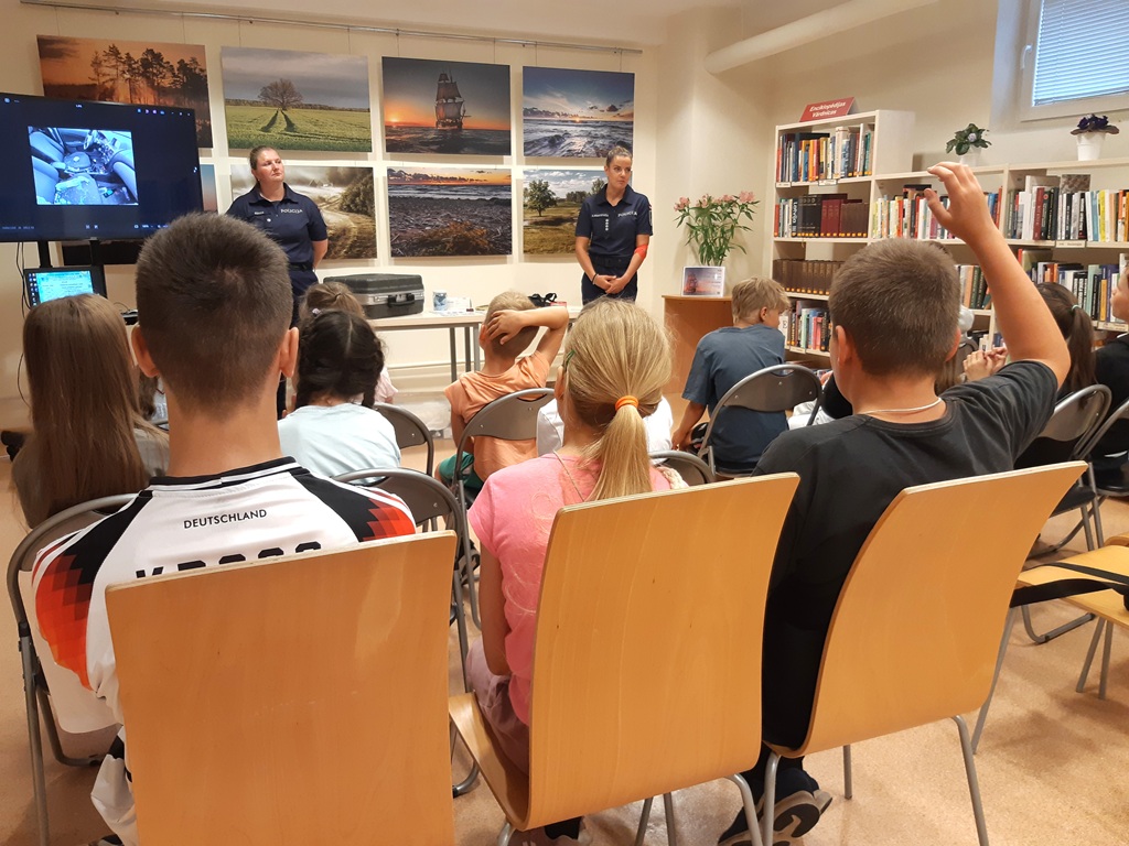 Divas policistes stāv telpas priekšā pie ekrāna, kamēr bērni sēž krēslos un viens no zēniem ceļ roku, lai uzdotu jautājumu.