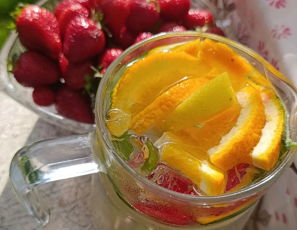 Stikla krūze ar atspirdzinošu dzērienu: apelsīnu un citronu šķēlītes, piparmētru lapiņas, ledus klucīši un ogas. Fonā – bļoda ar svaigām zemenēm.