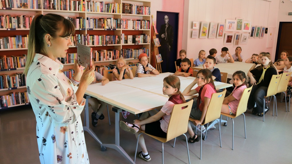 Bibliotekāre stāv pie galda un rāda grāmatu bērnu grupai, kas sēž un uzmanīgi klausās stāstītajā, fonā atrodas grāmatu plaukti un tālāk fonā izkārta izstāde.