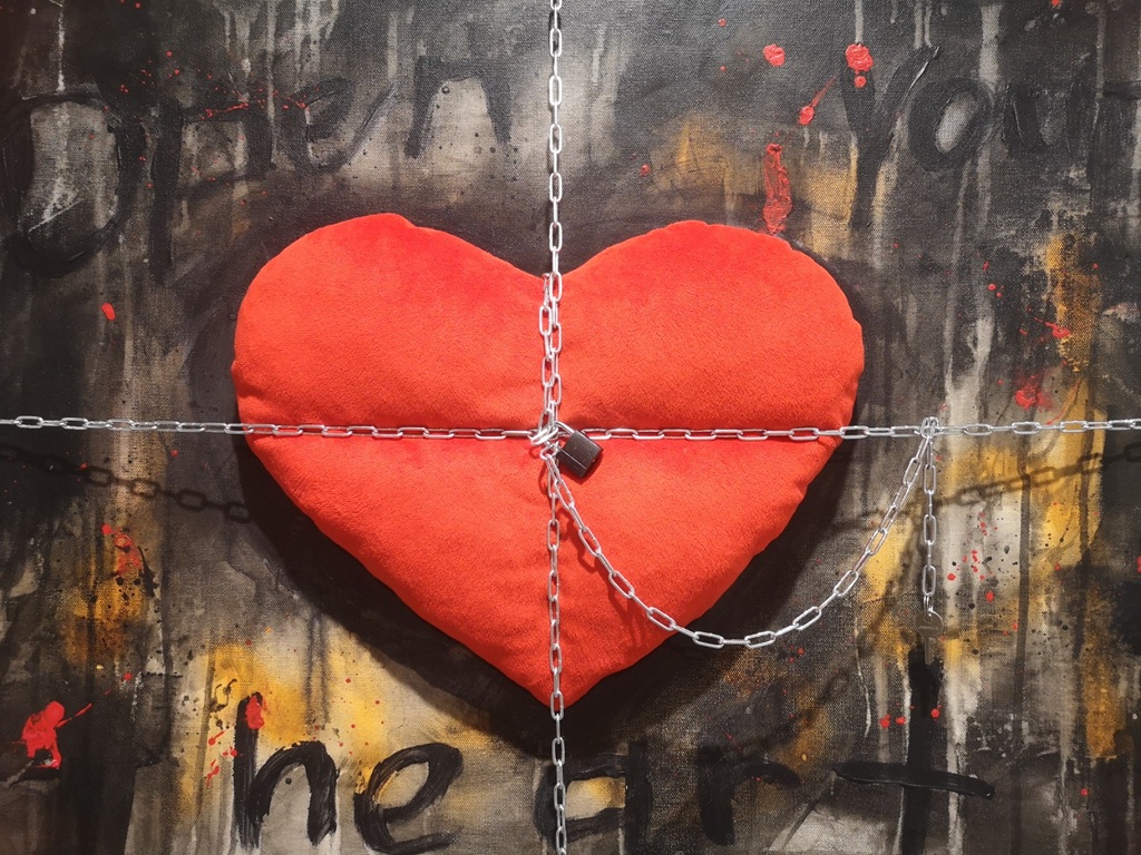 Žannas Vērdiņas mākslas darbs, kurā uz tumša, teksturēta fona ar abstraktiem triepieniem un daļēji saredzamu angļu valodas teikumu "Open your heart" centrā ir novietota liela, sarkana sirds formas mīkstā rotaļlieta. Sirds ir aptīta ar metāla ķēdēm, kas vidū saslēgtas ar piekaramo atslēgu, simbolizējot ierobežojumu vai noslēgtību.